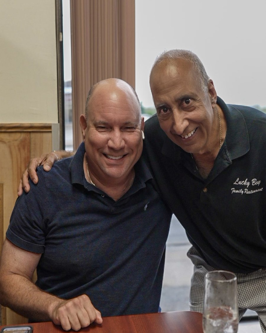 The owner of Lucky Boy Restaurant with his arm around a longtime regular, both smiling at a table in the dining room.