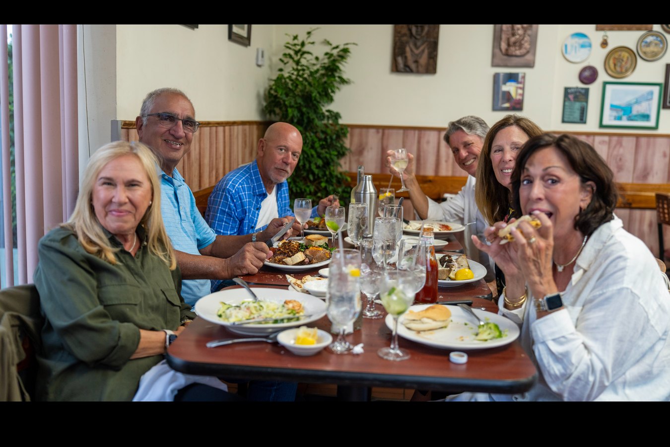 A group of six friends seated together at a table at Lucky Boy, mid-meal and laughing, plates and wine glasses in front of them.