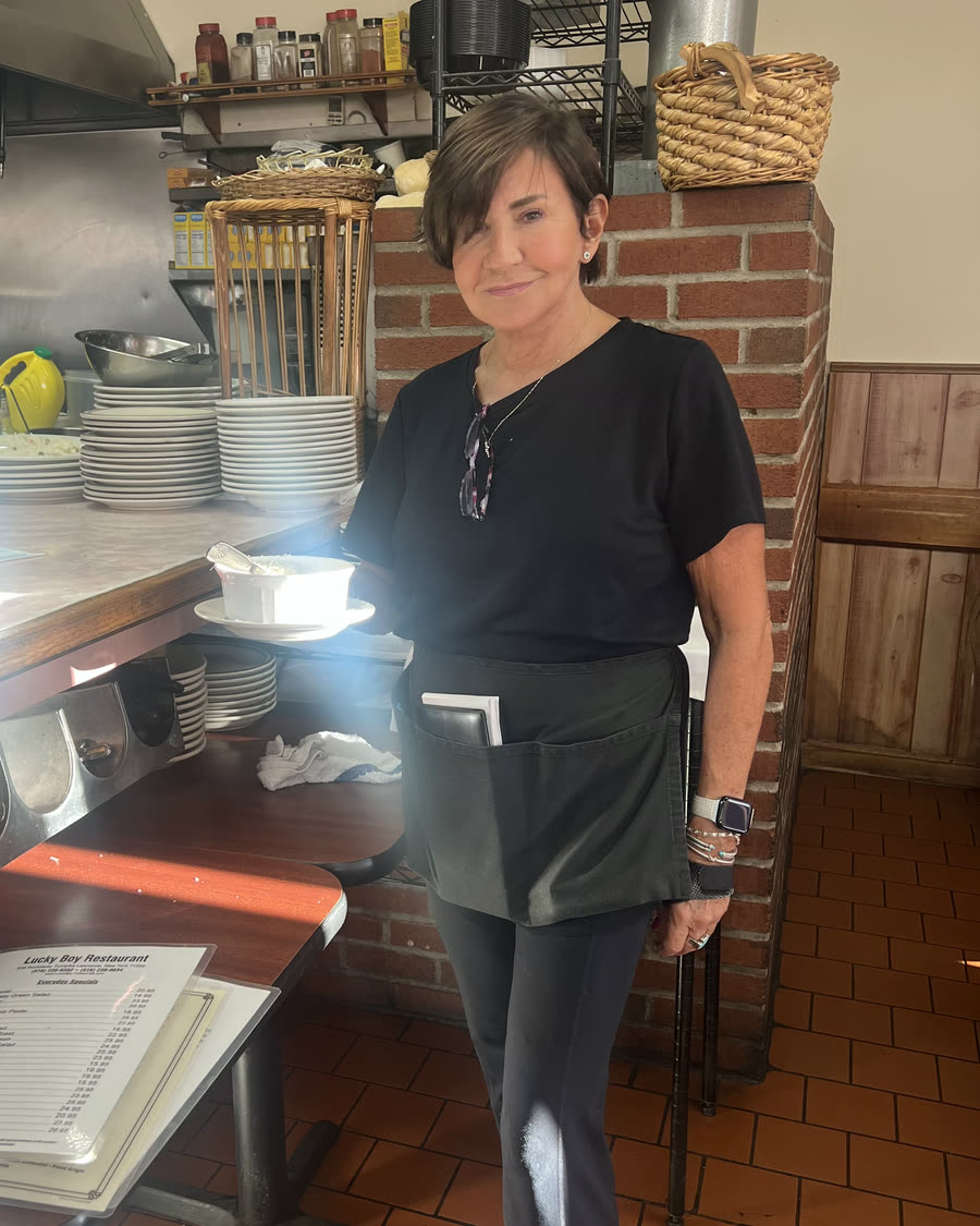 A longtime Lucky Boy server in a black shirt and apron, smiling beside the brick pillar near the counter, with stacks of plates behind her.
