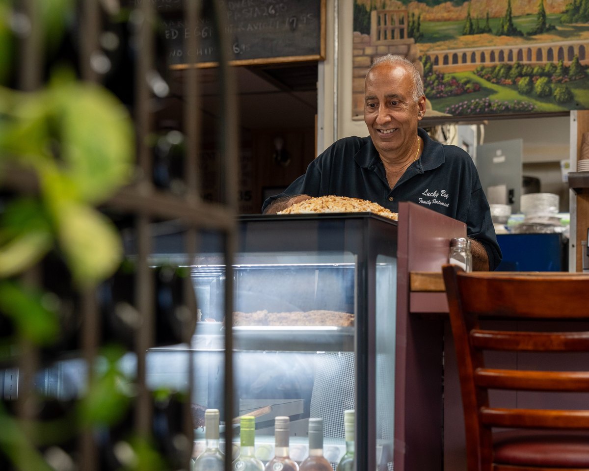The owner smiling behind the counter at Lucky Boy, beside the refrigerated dessert case.