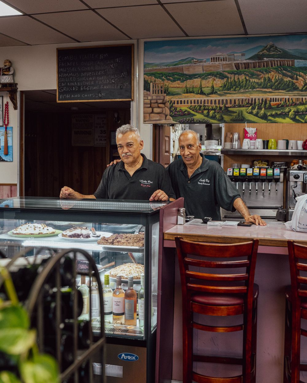 Two members of the Lucky Boy family standing together behind the counter, with the dessert case in front of them and a painted mural of the Acropolis on the wall behind.