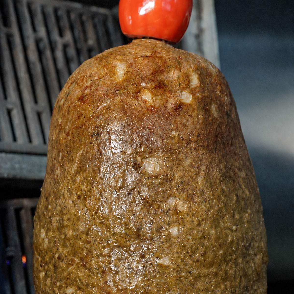 Gyro meat cone roasting on the vertical spit, topped with a whole tomato, ready to be carved.