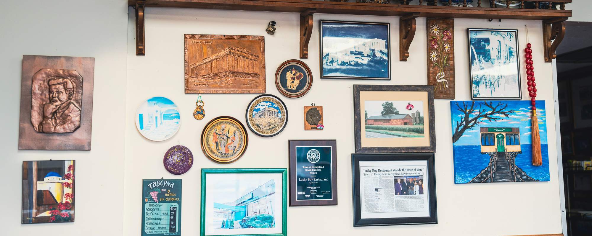 A wall inside Lucky Boy covered with Greek plates, copper reliefs and paintings, a shelf of Greek pottery above, and framed among them the Town of Hempstead Small Business Award and a newspaper article about the restaurant.
